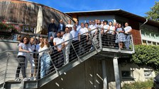 A group of people stands on an outdoor staircase in front of a building, posing for a photo. The sky is clear and blue.