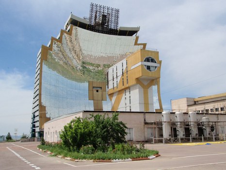 Ein großes Solarofengebäude mit einer spiegelnden, geschwungenen Fassade, einer rechteckigen Struktur an der Basis und Industrieanlagen auf der rechten Seite unter einem bewölkten Himmel.