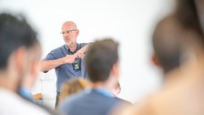 Im Vordergrund sind verschwommen die Hinterköpfe von Personen zu sehen. Scharf zu sehen ist eine Person mit Mikrofon um den Hals die gerade die Hände verschränkt.