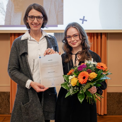 Two women stand smiling in front of a screen. The one on the left is holding a certificate and the one on the right a bouquet of flowers.