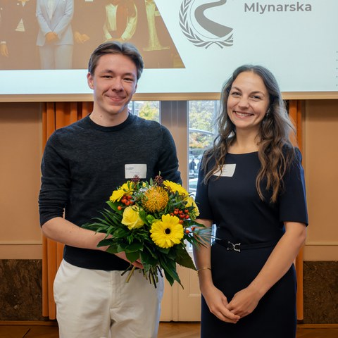 Ein Mann und eine Frau stehen vor einer Tür und einer Leinwand. Der Mann hält einen Blumenstrauß in den Händen. Beide lächeln.