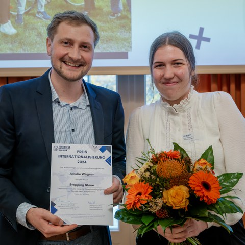 Ein Mann und eine Frau stehen vor einer Leinwand. Es sind nur ihre Oberkörper zu sehen. Der Mann hält eine Urkunde in den Händen und die Frau einen Blumenstrauß. Beide lächeln.