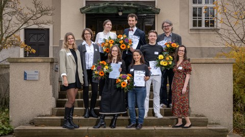 Eine Guppe von Menschen steht auf einer Treppe. Es ist ein Gruppenbild. Alle lächeln. Die sechs Personen in der Mitte halten alle einen Blumenstrauß und eine Urkunde in den Händen.