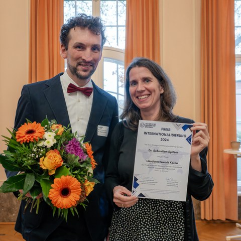 Ein Mann und eine Frau stehen vor einem Fenster mit orangenen Vorhängen. Der Mann hält einen Blumenstrauß in den Händen und die Frau präsentiert eine Urkunde. Beide Lächeln