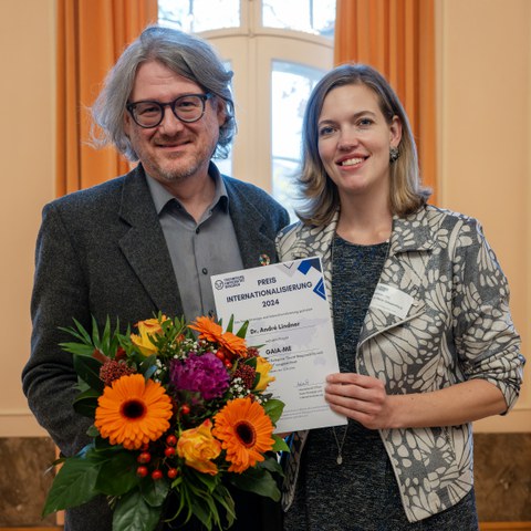 Ein Mann und eine Frau stehen vor einer Leinwand. Der Mann hält einen Blumenstrauß in den Händen und die Frau eine Urkunde