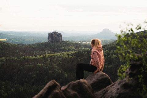 Frau in pinker Jacke sitzt auf einem Felsen. Im Hintergrund Wald und Felsformationen.