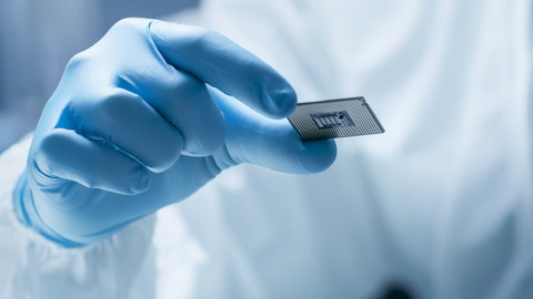 Bild einer Person in Handschuh und Laborkittel, die einen Mikrochip in die Kamera hält.