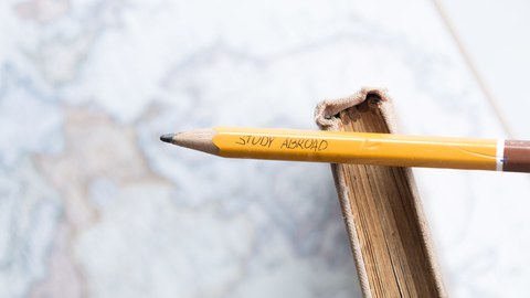 Foto eines Bleistiftes mit der Aufschrift "Study Abroad". Der Stift liegt auf einem Buch und beides befindet sich auf einer Weltkarte.