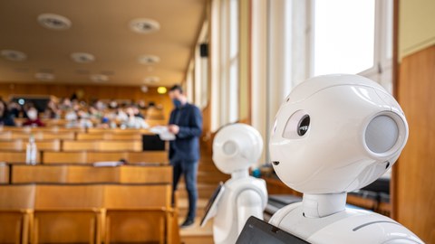 In the picture, two white robots can be seen in the foreground on the right-hand side. Both have a rectangular tablet in the chest area. The middle ground and background shows a lecture hall filled with students and is blurred. 