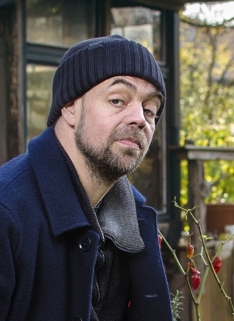 Porträtfoto von Sven Hönig. Er trägt eine dunkle Mütze und eine dunkle Jacke und sitzt im Freien, möglicherweise in einem Garten. Es ist Herbst.