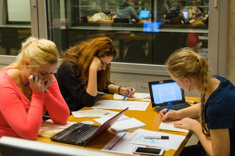 Drei Studentinnen arbeiten in einem Gruppenraum an einem Tisch, auf dem sie ihre Laptops und viele Handouts verteilt haben.