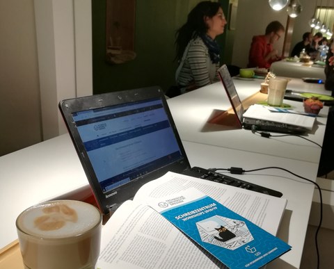 Das Café insgrüne als Arbeitsplatz für wissenschaftliches Schreiben: Zu sehen sind ein geöffneter Laptop sowie ein Flyer des Schreibzentrums, daneben eine Tasse Kaffee. Im Hintergrund die Reflexion von Cafébesuchern und Lampen.