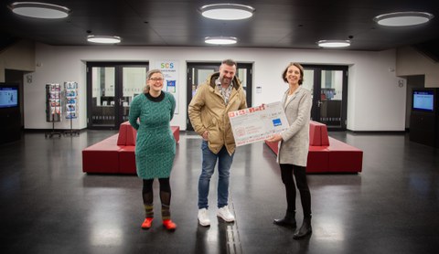 3 Personen stehen in einem Raum, die rechte Person hält einen großen Geldscheck in der Hand. Die Personen lachen.