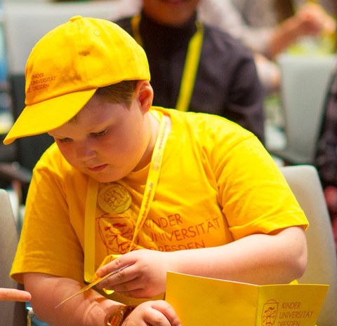 Ein Junge trägt ein gelbes Kinder-Uni-T-Shirt, ein gelbes Kinder-Uni-Basecap auf dem Kopf und ein gelbes Kinder-Uni-Schreibheft in der Hand.
