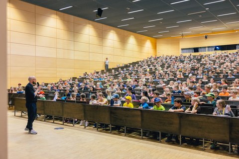 Das Foto zeigt einen Hörsaal, der mit Kindern gefüllt ist. Vor den Sitzreihen steht eine männlich gelesene Person, die in ein Mikrofon spricht und in Richtung der Sitzreihen guckt.