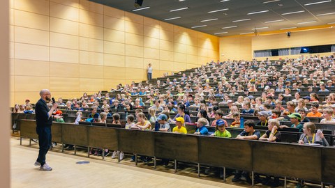 Das Foto zeigt einen Hörsaal, der mit Kindern gefüllt ist. Vor den Sitzreihen steht eine männlich gelesene Person, die in ein Mikrofon spricht und in Richtung der Sitzreihen guckt.