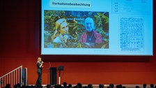 Referentin steht vor einer Menge Publikum, der Beamer zeigt eine Folie mit der Frage Wie erforschen wir, was Affen denken? und 2 Fotos von Jane Goodall sowie Aufzeichnungen einer wissenschaftlichen Studie zum Verhalten von Affen