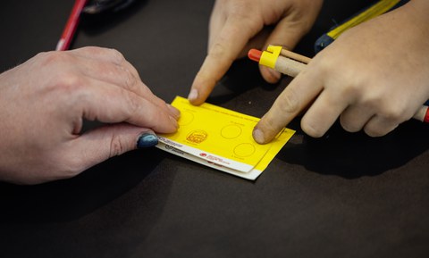Kinder-Uni-Ausweis mit einem Stempel, 3 Hände sind zu sehen, 1 Hand hält einen Kuli