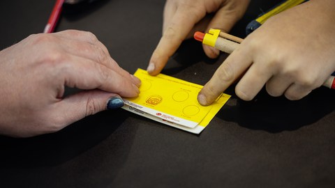 Kinder-Uni-Ausweis mit einem Stempel, 3 Hände sind zu sehen, 1 Hand hält einen Kuli