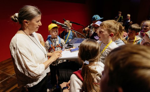 Eine Frau steht an einem Rednerpult. Ihr gegenüber steht ein Junge, der mit ihr spricht. Um die beiden herum stehen weitere Kinder.