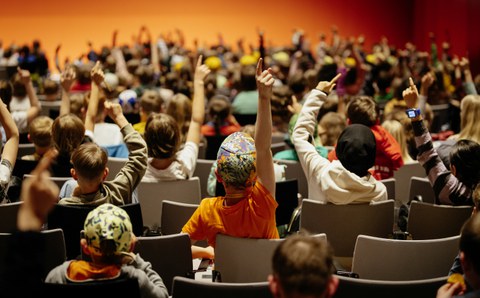Viele Kinder melden sich und recken die Arme in die Höhe. Sie sitzen in Reihen und sind von der Rückseite zu sehen.