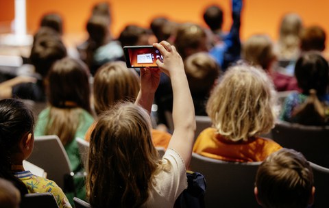 Ein Kind ist von hinten zu sehen. Es sitzt auf einem Stuhl zwischen anderen Kindern und hält ein Smartphone in die Höhe. Damit fotografiert das Kind etwas.