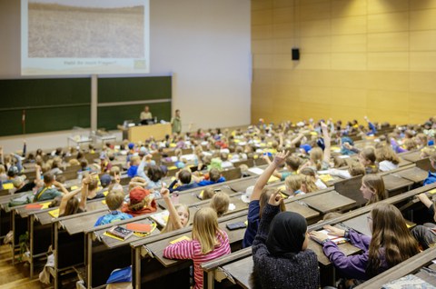 Viele Kinder sitzen in einem Hörsaal und melden sich. Dieses Bild zeigt die Kinder von hinten.