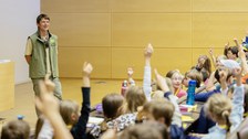 Viele Kinder sitzen in einem Hörsaal und melden sich. Vor ihnen steht ein Mann und sieht lächeln zu ihnen.