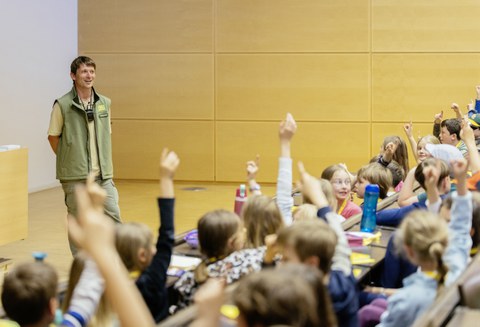 Viele Kinder sitzen in einem Hörsaal und melden sich. Vor ihnen steht ein Mann und sieht lächeln zu ihnen.