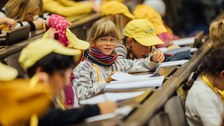 Kinder sitzen in den Sitzreihen eines Hörsaals und machen Notizen.