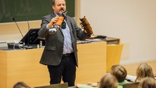 Ein Mann steht vor einer Tafel und einem Pult in einem Hörsaal und hält zwei Stofftiere in der Hand.