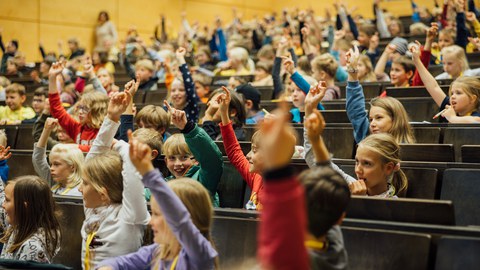 Viele Kinder heben die Hände und stellen Fragen.