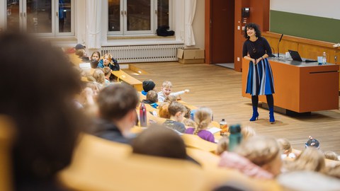Das Foto zeigt eine Szene in einem Hörsaal. Vor der Tafel steht eine Referentin und spricht zu den Kindern, die in den Reihen sitzen.