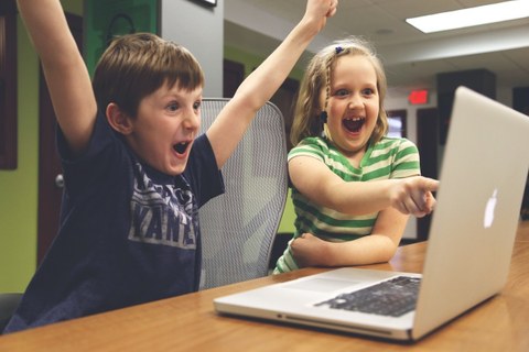 Ein Junge und ein Mädchen sitzen am Tisch vor einem aufgeklappten Laptop. Beide jubeln.