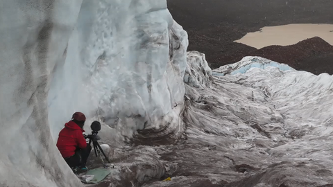 Der Screenshot von der Vorlesung von Jun.-Prof. Dr. Miriam Akkermann zeigt Frau Akkermann auf einem Gletscher. Sie befindet sich links im Bild. Vor ihr steht ein Mikrofon.