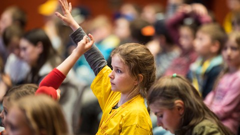 Das Foto zeigt ein Mädchen im Hörsaal, das seine Hand hebt. Das Mädchen trägt ein T-Shirt der Kinder-Uni.