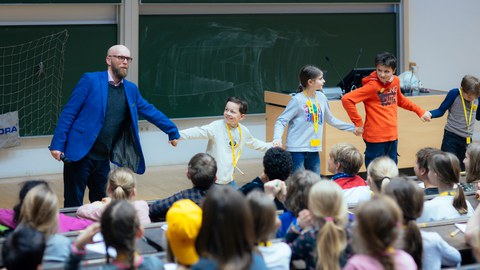 Das Foto zeigt den Moderatoren und vier Kinder, die sich an den Händen halten und ziehen. Sie veranschaulichen damit bei einem Experiment, was der Referent gesagt hat. 