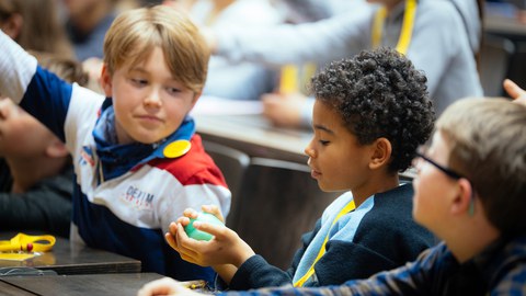 Das Foto zeigt drei Jungen im Hörsaal. Der Junge in der Mitte hat einen kleinen grünen Ball in der Hand. Den Ball hat der Professor den Kindern zur Veranschaulichung gegeben. 