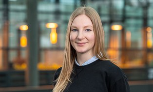 Das Bild zeigt ein Portraitfoto von einer blonden Frau, die einen schwarzen Pullover trägt. Der Hintergrund ist verschwommen, es sind eine Glasfront und orange Lichter zu erkennen.