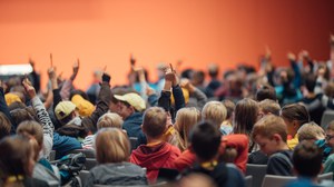 Das Foto zeigt Kinder, die im Saal sitzen. Viele von den Kindern heben die Hand.  