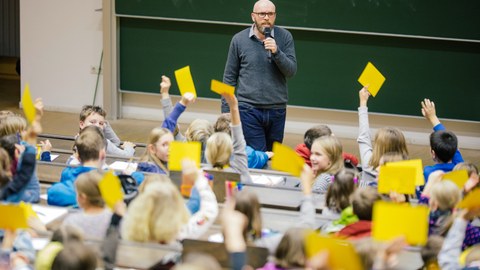 Das Foto zeigt Kinder im Hörsaal von hinten, die gelbe Karten hochhalten. Vorn im Saal steht der Moderator und spricht zu den Kindern. 