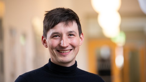 Das Foto zeigt das Portrait eines Mannes, der braunes Haar hat und einen schwarzen Pullover trägt.