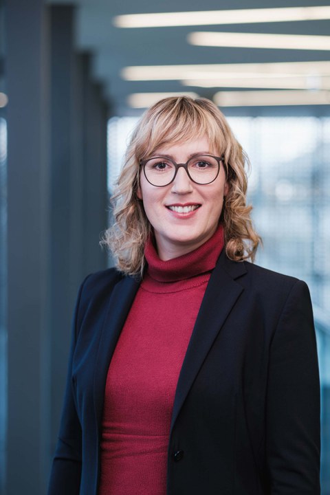 Das Foto zeigt das Portrait einer weiblich gelesenen Person, die eine Brille und schicke Kleidung trägt und in die Kamera lächelt.