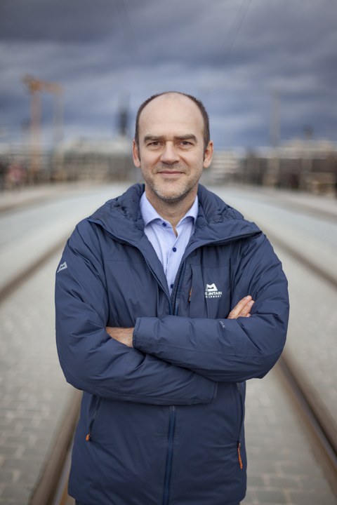 Das Foto zeigt einen Mann, der auf einer Straße steht. Er hat die Arme verschränkt und lächelt in die Kamera.