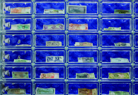 Das Foto zeigt eine blaue Wand, vor der Glasbausteine stehen. In jeden Glasbaustein liegt ein Geldschau unterschiedlicher Höhe und Herkunft.