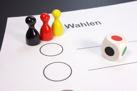 Das Bild zeigt einen Teil eines Abstimmungszettels mit der Überschrift Wahlen. Es sind 3 Spielfiguren in schwarz, rot und gelb und ein Spielwürfel zu sehen.