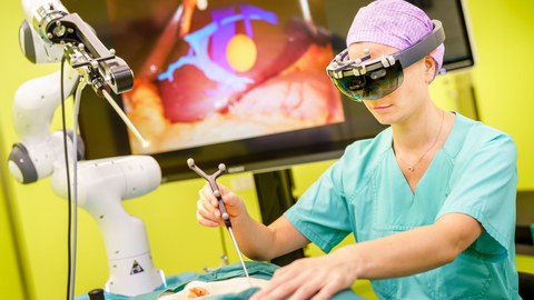 Eine Frau steht an einem OP-Tisch und hat ein OP-Instrument in der Hand. Im Hintergrund ist ein Bildschirm zu sehen. 