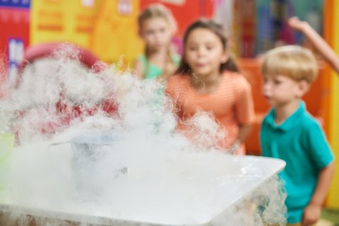 Auf einem Tisch im Vordergrund steht ein Gefäß, aus dem Nebel kommt. Im Hintergrund sind Kinder, die den Nebel beobachten.