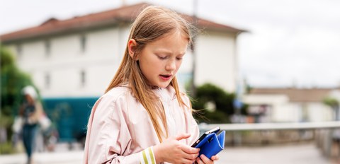 Mädchen mit Handy in der Hand, der Mund ist leicht geöffnet. Im Hintergrund ein großes Gebäude und weitere Personen.