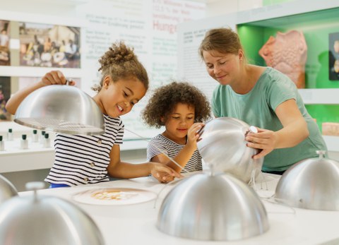 Das Bild zeigt zwei Kinder und eine erwachsene Person. Sie stehen an einem Tisch, auf dem Tisch sind Abdeckhauben. Sie gucken unter zwei der Abdeckhauben. Im Hintergrund ist eine Ausstellung.